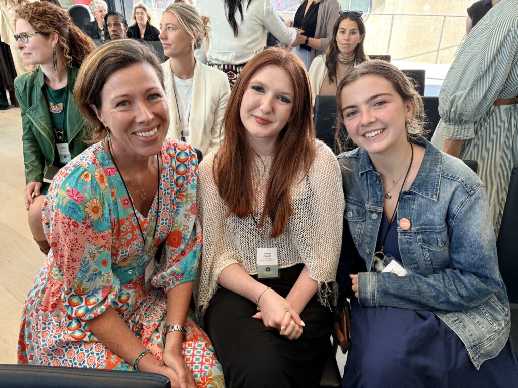 Three smiling women seated together at a conference, left in a floral dress, middle with red hair, right in a denim jacket with a badge visible.